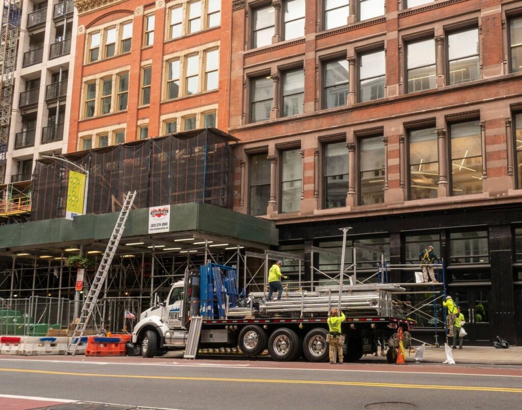 NYC renovation project with construction crew setting up scaffolding and safety barriers in front of a commercial building, indicating active work under city permit.