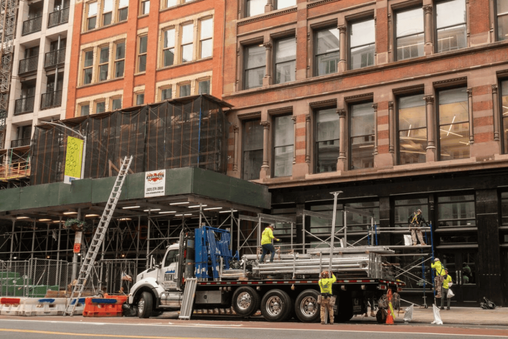 NYC renovation project with construction crew setting up scaffolding and safety barriers in front of a commercial building, indicating active work under city permit.